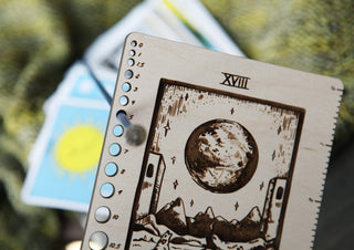 A close-up of the 🌙 The Moon 🌙 Tarot Card Knitting Needle Gauge & Ruler, labeled XVIII, with its unique needle gauge scoring system. Blurred tarot cards and green fabric appear in the background.