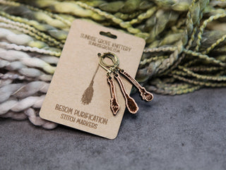 A set of 🧹 Witch’s Broom Stitch Markers 🧹 rests on green and cream chunky yarn, ready to add magical charm to your knitting or crochet projects.