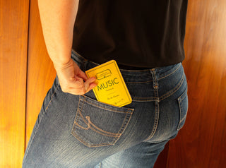 A person wearing blue jeans and a black shirt places a Pocket Music Notebook with a yellow cover into their back pocket. Wooden panels can be seen in the background.
