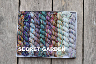 A Koigu Pencil Box in the Secret Garden colorway, filled with fingering-weight yarn and arranged in neat rows on a wooden surface. The words Secret Garden appear in bold white letters at the bottom left.