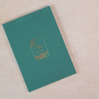 The Calligraphy Practice Pad, in green A5 size, features a gold embossed illustration of a house, trees, and birds on the cover, and is displayed on a light wooden surface.