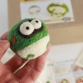 A hand holds a small, round felted wool frog with big eyes in front of a blurred DIY Needle Felting Kit – Frog and instructions—great for anyone starting with a beginner DIY needle felting kit.