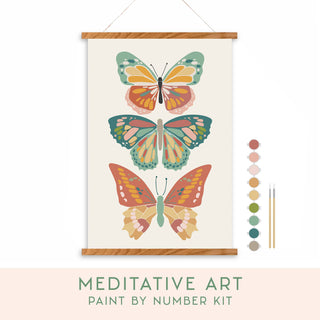 The Vibrant Butterflies Meditative Art Paint by Number Kit features three stylized butterflies in earthy tones, shown vertically. On the right are six paint colors and three brushes. Below, text reads: "MEDITATIVE ART PAINT BY NUMBER KIT.