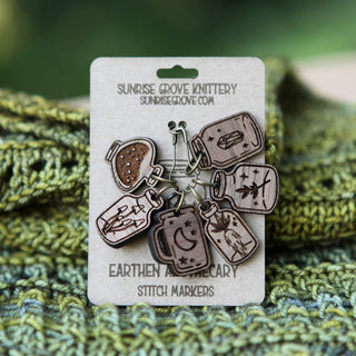 The Apothecary Bottle Stitch Marker Set includes six walnut wood markers, each engraved with a unique apothecary bottle design, displayed on a branded card—ideal knitting accessories for any maker.