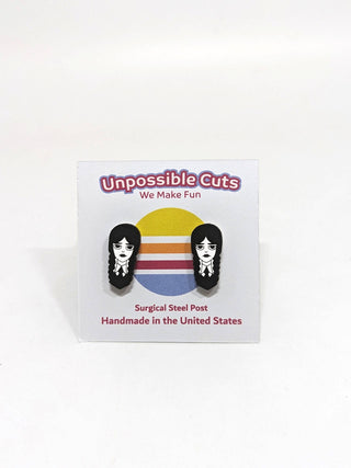 A pair of black and white Wednesday Stud Earrings featuring a cartoon girl with braids, hypoallergenic and handmade in San Francisco from sustainable wood, displayed on an Unpossible Cuts—We Make Fun card with a retro striped circle.