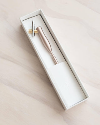 The Flourish Calligraphy Pen, featuring a pointed nib and sleek handle ideal for beginners, rests in a white rectangular box on a light wooden surface.