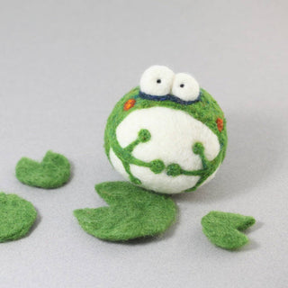 A round, green and white felt frog with bulging eyes sits on a gray surface among green lily pads—a perfect beginner project with the DIY Needle Felting Kit – Frog.