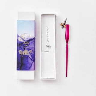 The Flourish Calligraphy Pen, with a metallic pink handle and attached nib, rests beside an open white box with a purple lid. Inside the box are a small tree illustration and a handwritten note.