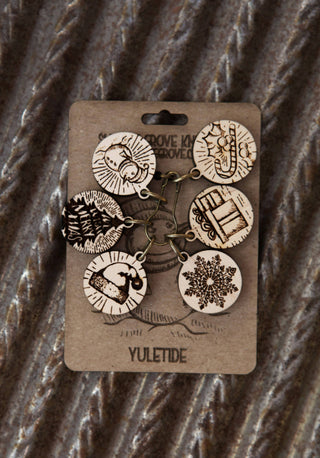 The Maple Wood Yuletide Knitting Stitch Markers Set of 6 features round ornaments with engraved festive designs, displayed on a brown YULETIDE card against a fabric background—ideal for adding holiday charm to your knitting projects.