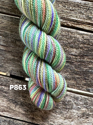 A skein from the KPM and KPPPM Variegated Color Collection, featuring green, purple, orange, and cream hues, rests on a rustic wooden surface. The label "P863" is displayed in white beside the yarn.
