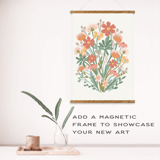 A Wildflowers Meditative Art Paint by Number Kit painting in a magnetic wooden frame hangs above a wooden block and a glass vase with greenery. Text: Add a magnetic frame to showcase your Wildflowers mindful painting.