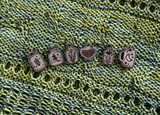 The Apothecary Bottle Stitch Marker Set features six small markers engraved with unique designs, displayed on a green, yellow, and black knitted fabric—ideal accessories for any knitting project.