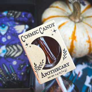 A close-up of a lollipop from the Season of the Witch Curio Box in moon-themed packaging, held before a white pumpkin with orange stripes and a purple cloth accented with blue and white patterns.