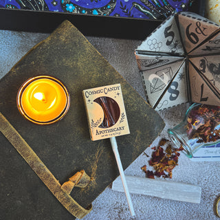 Beeswax candle next to a lollipop in 'Cosmic Candy Apothecary' packaging on a textured surface with other items.
