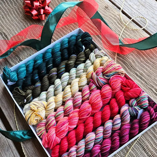 The Koigu Pencil Box in "A Christmas Story" features colorful mini skeins of Merino Wool Yarn, arranged from deep green to reds and variegated shades. Red and green ribbon, gold string, and a red bow accent the rustic wooden surface.