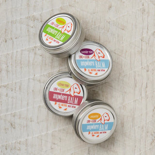 Four small tins of Little Bee Anywhere Balm—a moisturizing beeswax balm—come in four scents: lavender-mint, citrus-rose, unscented, and orange-lemongrass, displayed on a light textured surface.