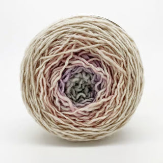 A Freia Ombre & Gradient Sport Wool Yarn cake shows a gradient from light cream on the outer edge to soft lavender and pale gray at the center, set against a plain white background.
