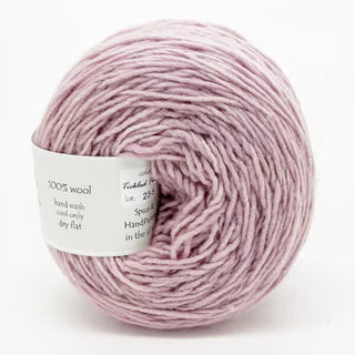 A ball of Freia Semi-Solid Sport Wool Yarn in light purple, heavy sport weight, with a white label detailing material and care. Made from 100% US wool, it looks soft and loosely spun.