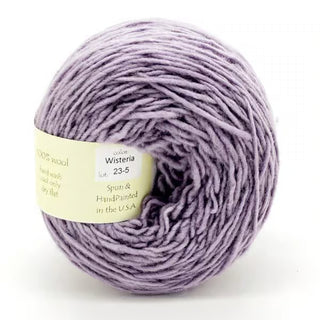 A skein of Freia Semi-Solid Sport Wool Yarn in soft, light purple Wisteria is wound in a ball with a cream label. This sport weight yarn has a smooth, slightly fluffy texture—ideal for cozy projects.