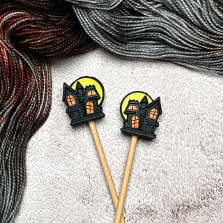 Knitting Needle Stitch Stoppers—wooden sticks with rubber haunted house decorations, set on a textured white surface with gray and reddish-brown yarn—make whimsical, practical knitting accessories.