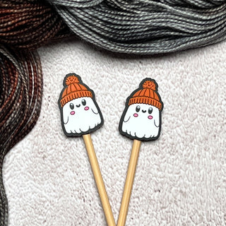 Knitting Needle Stitch Stoppers—two wooden needles with adorable ghost-shaped silicone stoppers in orange knit hats, set on a textured beige surface with yarn accents—make charming, fun knitting accessories.