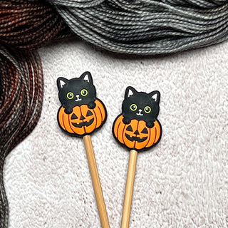 Knitting Needle Stitch Stoppers featuring black cat faces in orange jack-o’-lantern silicone design on needles, displayed on textured white surface with gray and brown yarn—ideal Halloween-themed knitting accessories.