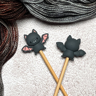 Knitting Needle Stitch Stoppers: A pair of wooden knitting needles with black bat silicone stoppers—one shows a smiling face and pink wings, the other its back. Resting on textured fabric with dark and grey yarn, these make charming knitting accessories.