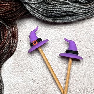 Two bamboo knitting needles with purple Knitting Needle Stitch Stoppers shaped like witch hats—one featuring an orange buckle—rest on a textured cream surface, with black, gray, and brown yarn in the background.