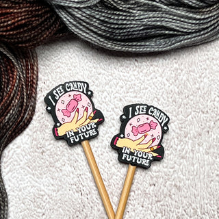 Two Knitting Needle Stitch Stoppers with pins that read "I SEE CANDY IN YOUR FUTURE" and display candy-filled crystal balls are placed on a textured white surface, surrounded by skeins of gray and brown yarn.