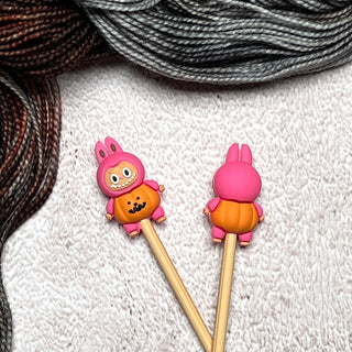 Two Knitting Needle Stitch Stoppers shaped like pink and orange cartoon bunnies rest on a textured white surface, with brown and gray yarn in the background—perfect for bringing charm to your knitting accessories collection.