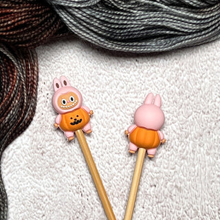 Two knitting needles with Knitting Needle Stitch Stoppers shaped like pink bunnies in orange pumpkin outfits rest on white textured fabric, with gray and brown yarn skeins in the background—ideal for your knitting accessories collection.