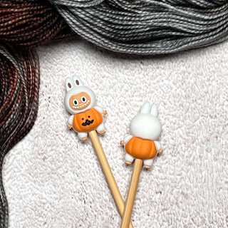 Knitting Needle Stitch Stoppers featuring cute bunny figures in orange pumpkin outfits on the ends of two chopsticks, displayed on a white textured surface with black, gray, and brown yarn—fun and charming knitting accessories.