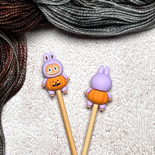 Knitting Needle Stitch Stoppers with cute animal toppers in purple and orange outfits, shown next to brown and gray yarn—fun, functional knitting accessories perfect for any project.
