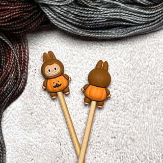 Two Knitting Needle Stitch Stoppers with cute brown pumpkin animal toppers are shown on a textured white surface, with gray and brown yarn skeins in the background.