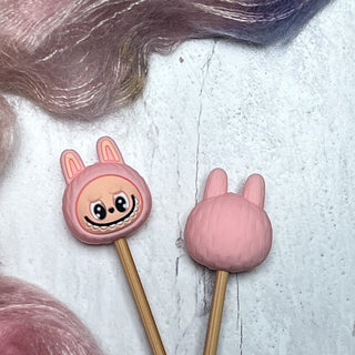 Two pink bunny-shaped Knitting Needle Stitch Stoppers are shown—one with a smiling cartoon face, the other with a textured back—amid wispy pastel fibers. These whimsical accessories keep your stitches secure while you knit.