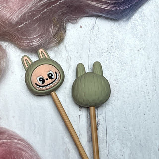 Knitting Needle Stitch Stoppers feature two needles with cute cartoon animal tops in green hoods with bunny ears—one smiling, one showing its back. Pink and purple yarn add a playful background to this fun knitting accessory set.