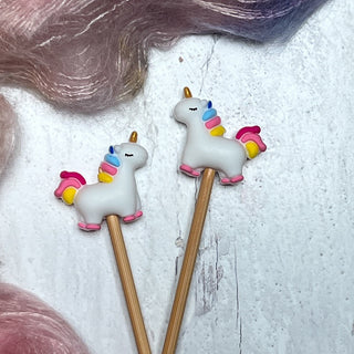 Two wooden sticks topped with small unicorns featuring colorful manes—these whimsical Knitting Needle Stitch Stoppers are perfect accessories for your knitting projects.