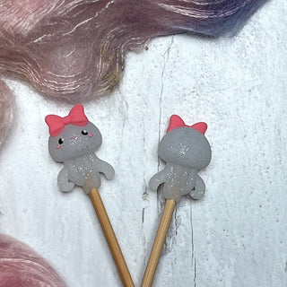 Two Knitting Needle Stitch Stoppers topped with cute gray octopus figurines, each featuring a pink bow and glittery finish, rest on a white wooden surface near pink and purple fibers—fun accessories for any knitting project.