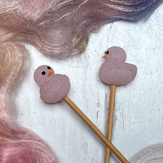 Two pastel pink Knitting Needle Stitch Stoppers shaped like ducks with orange beaks rest on wooden sticks, displayed on a white surface decorated with wispy, iridescent multicolored fibers.