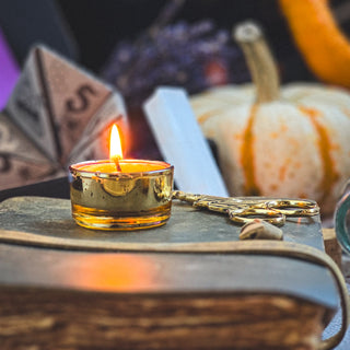 A small gold candle burns on a closed, old book next to ornate scissors. In the background, a white pumpkin and autumn decor create cozy vibes—perfect for mystical knitters. Discover it all in the Season of the Witch Curio Box.