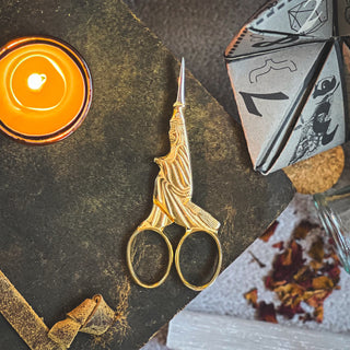 A pair of ornate gold scissors shaped like a robed figure rests on a textured dark surface, accompanied by mystical items from the Season of the Witch Curio Box—a lit orange candle, folded paper fortune teller, dried leaves, and other enchanting gifts.