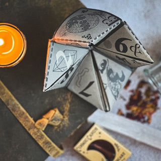A paper fortune teller with witchy designs rests on a table near a lit candle, dried petals, a strapped vintage book, and the Season of the Witch Curio Box, creating a magical scene.