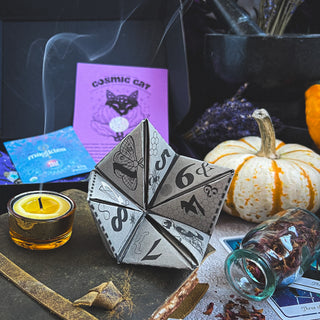 A mystical paper die with symbols rests amid tarot cards, a white pumpkin, herbs, a tealight, and purple cards—an enchanting scene inspired by the Season of the Witch Curio Box, perfect for magical knitting gifts.