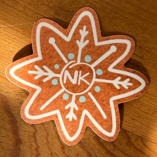 The Knitmas Stickers feature a gingerbread-shaped premium vinyl sticker with white and light blue snowflake icing and "NK" in the center, displayed on a wooden surface in warm sunlight—the perfect festive touch.