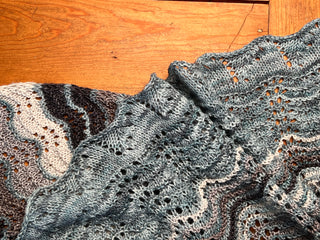 A close-up of a rectangle shawl crafted with wavy, lacy Old Shale motif patterns in shades of blue, gray, and cream, resting on a wooden surface—perfect for your next project using the Knitmas in July 2026 Pattern.