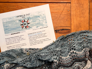 A printed knitting pattern titled Knitmas in July 2026 Pattern sits on a wooden table next to a partially completed blue and gray lace knit project with stitch markers attached—a beautiful rectangular shawl featuring the classic Old Shale motif for the Knitmas in July Knit-Along.