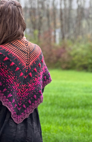 A triangular Season of the Witch 2026 Shawl Pattern featuring a dark grey base with a gradient yarn pattern of orange, pink, and purple textured stitches.