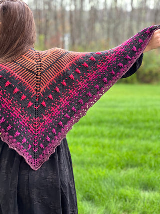A person wearing a gothic lace Season of the Witch 2026 Shawl Pattern, featuring textured stitches that fade from orange at the top to pink and purple at the edges against a black background.