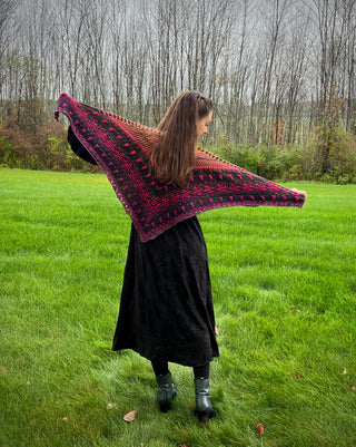 A person in a black dress stands in a grassy field, holding out a Season of the Witch 2026 Shawl Pattern featuring a gradient of black, purple, and orange geometric patterns.