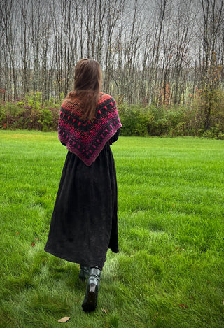 A person seen from behind wears a black dress and a Season of the Witch 2026 Shawl Pattern featuring a gradient of black, purple, and orange geometric patterns, standing in a grassy field in front of a forest.
